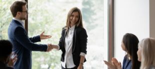 Two coworkers shake hands in a bright conference room while colleagues applaud in the background.