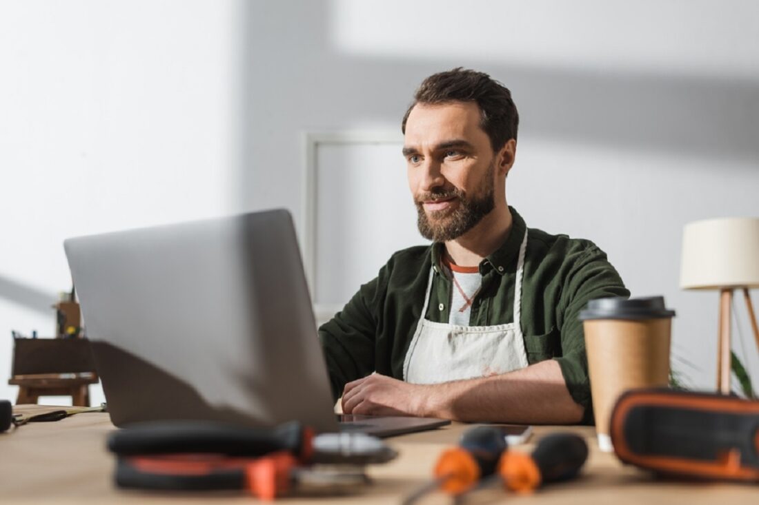 Bearded,Carpenter,Using,Laptop,Near,Blurred,Takeaway,Coffee,And,Tools
