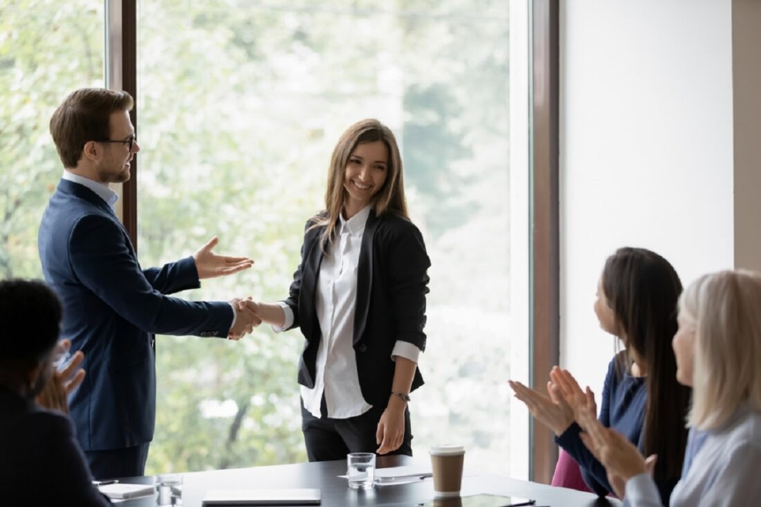 Two coworkers shake hands in a bright conference room while colleagues applaud in the background.