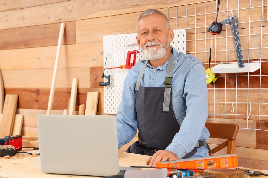 Mature,Carpenter,Working,With,Laptop,At,Table,In,Shop