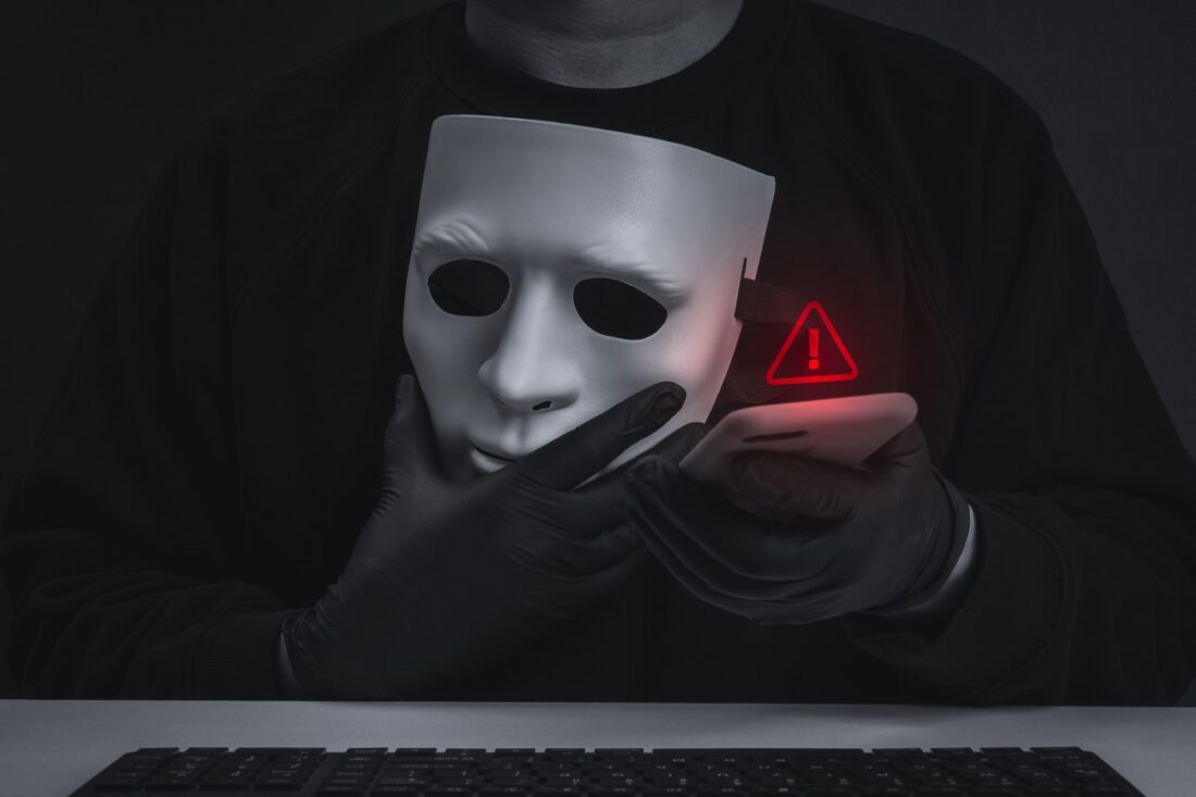 Dark,Conceptual,Image,Of,Masked,Cybercriminal,Scammer,Holding,Smartphone,Beside